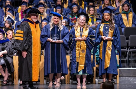 Dean Stephen Cavanagh, left, stands next to three graduates holding crystal awards on commencement stage, all wearing cap and gown
