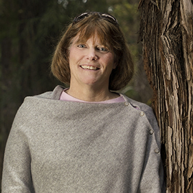 A woman wearing a gray sweater stands next to a redwood tree.