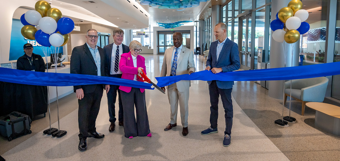 5 hospital leaders stand inside new center each holding part of a blue ribbon while center woman uses large scissors to cut it
