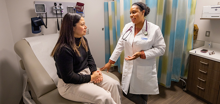 A doctor in a white coat, right, gestures with her left hand while speaking with a patient who is seated on an exam room chair
