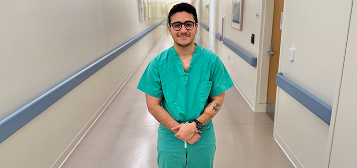 Medical student with dark hair, eyeglasses, green scrubs and tattoo on his left arm stands with hands crossed in an empty hallway at UC Davis Medical 