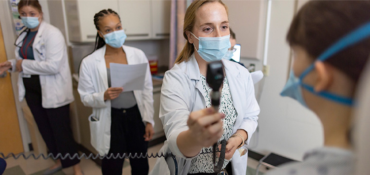 Student wearing mask and white coat extends arm holding ophthalmoscope up to patient’s eyes while other students look on