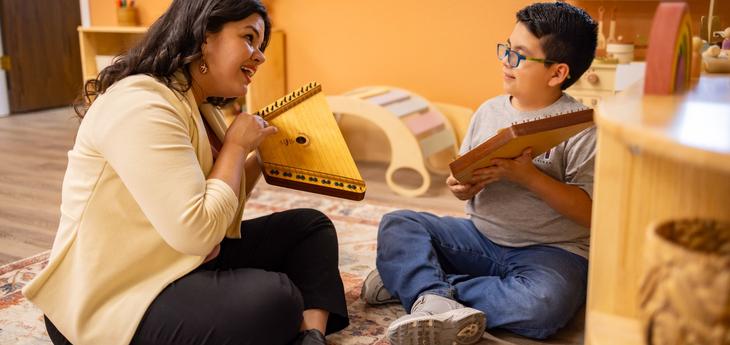 A woman and a young boy sit on the floor in a warm, inviting classroom, smiling and playing wooden string instruments together.