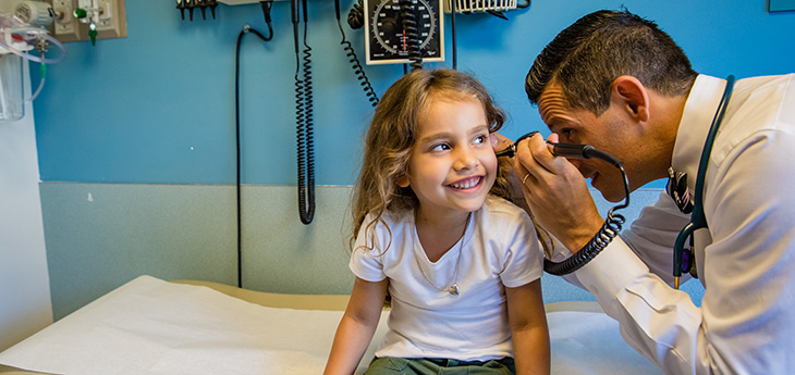 Doctor looks in girl’s ear while she smiles on patient table.  