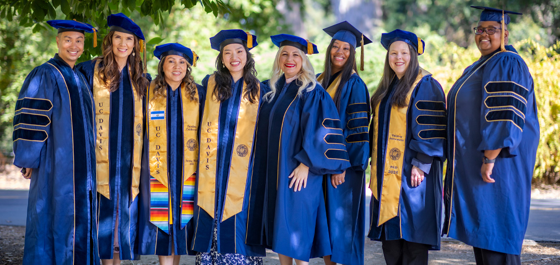 Eight graduates in commencement cap and gowns stand next to each other outside and smile at camera