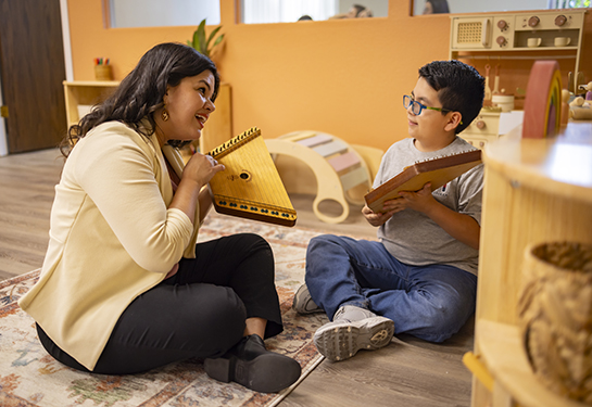 A woman and a young boy sit on the floor in a warm, inviting classroom, smiling and playing wooden string instruments together.