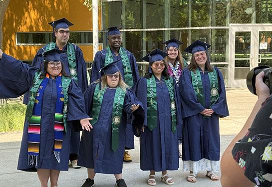 A group of seven Redwood SEED scholars in navy blue caps and gowns and green stoles celebrate their graduation. 