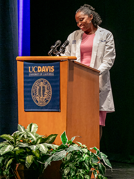 A doctor in white coat speaks at the podium, smiling as she looks down at her notes