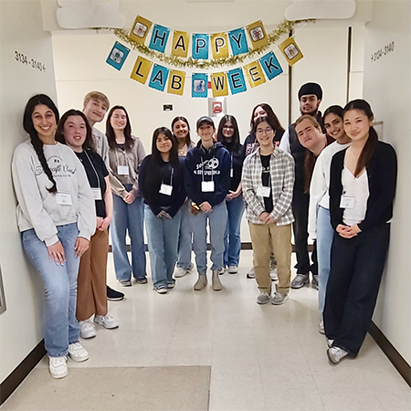 Fourteen high school students stand in a hallway with an overhead sign that reads: “Happy Lab Week.”