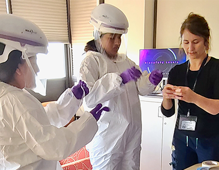 Two high school students dressed in full-body protective suits with helmets, face shields, and purple gloves.