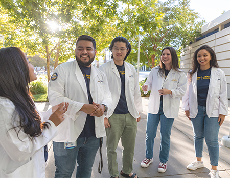 Five medical students in white lab coats and jeans converse outside the Education Building at UC Davis School of Medicine