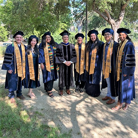A doctor in black gown poses with seven students in graduation garb at the UC Davis medical school’s commencement on May 17, 2025.