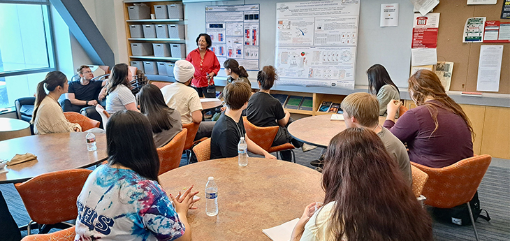 A woman in a red blouse stands at the front of a classroom before about 15 high school students seated around tables