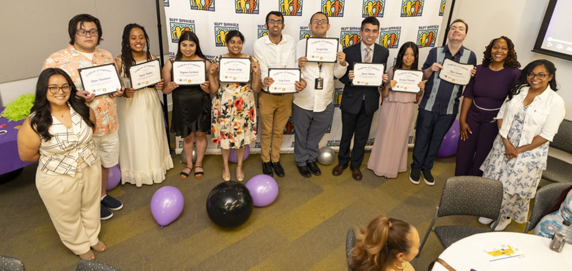 A group of people holding certificates pose in front of a large panel that reads "Best Buddies"