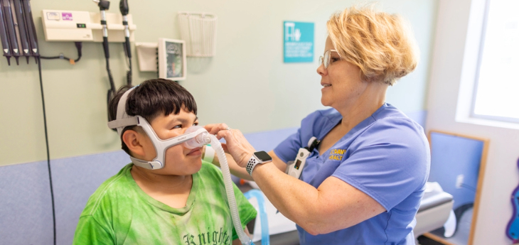 Child with green shirt being fitted with oxygen mask by respiratory therapist. 