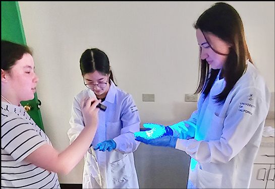 Three high school students, two of them in white lab coats, use a blue flashlight to learn about germs and hand hygiene 