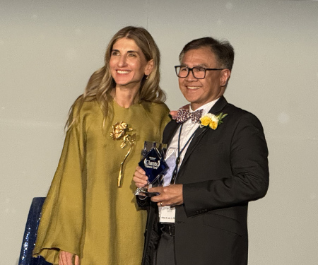 Woman with brown hair wearing long flowing gold-colored gown next to man in dark suit and brown polka dot tie holding trophy.