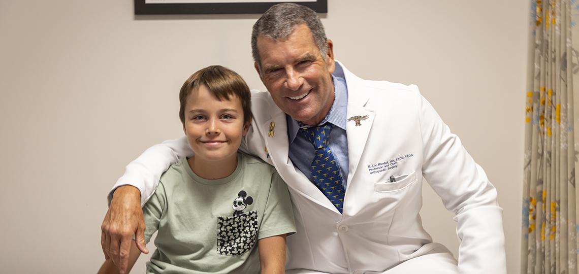 Doctor in a white coat with his arm around a young boy with a green shirt sitting on an exam table.