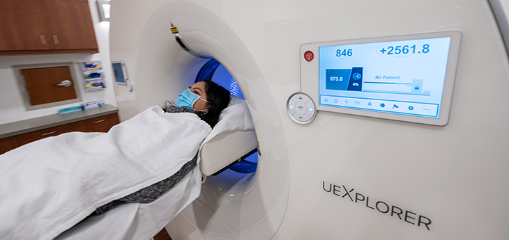 Patient with dark hair wearing a mask lying down at entrance to large cylindrical machine with the words UEXPLORER written on the outside.