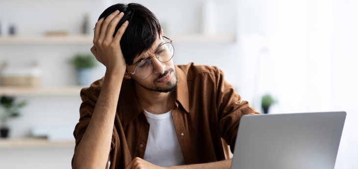 Exhausted man sitting in front of laptop at office, touching his head.