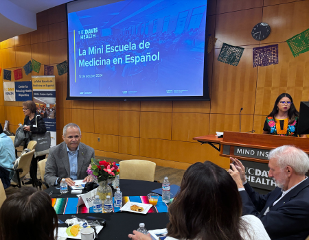 People sit around tables in a room where a projection screen shows a slide that states, “La Mini Escuela de Medicina en Español”