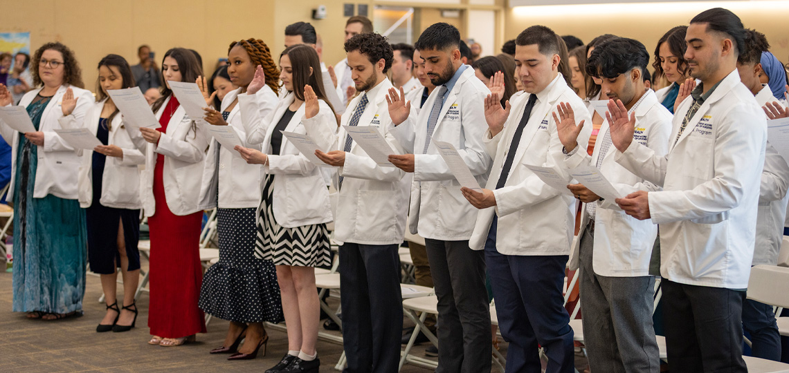 A group of students wearing white coats stand in a row, holding papers and raising their right hands as they take an oath during a ceremony.