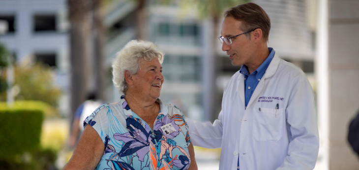A person in a white coat speaking with someone in a floral shirt using a walker.