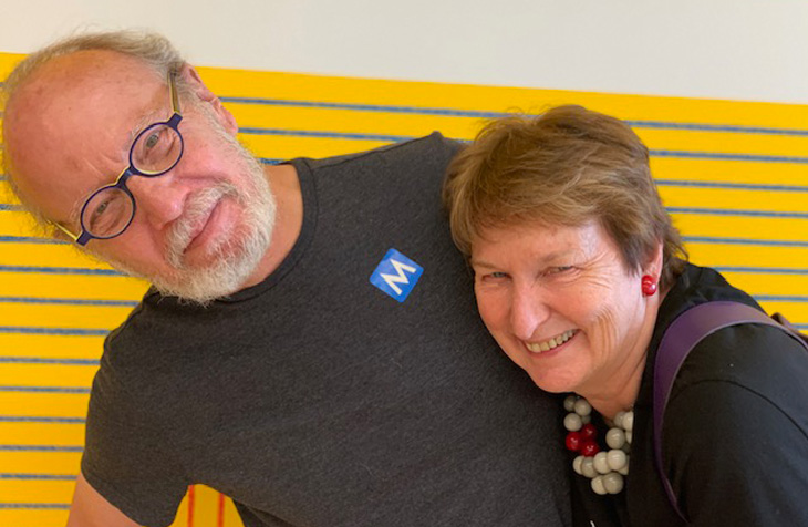 Two people smiling and leaning together in front of a yellow wall with blue stripes.