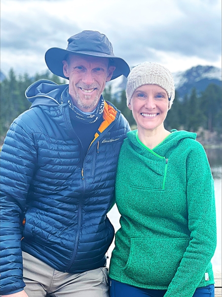 A smiling man and woman stand in a nature setting with trees and mountains in the background.
