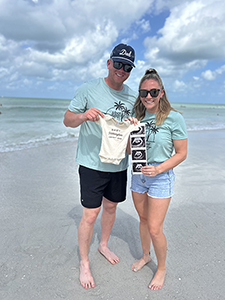 Man and woman standing on a beach holding a white infant onesie an ultrasound with four images of a developing baby.