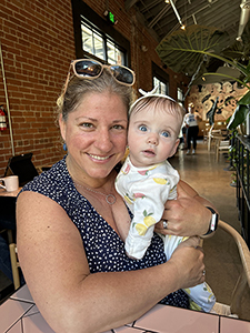 Blonde woman with sunglasses on her head holds a little baby girl in a white onesie with fruit on it and a white headband with a bow.