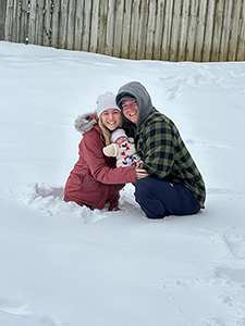 Woman in mauve colored jacket with fur hood and white knit cap with a man in a black and grey checkered with grey hood both holding an infant while wait deep in snow.
