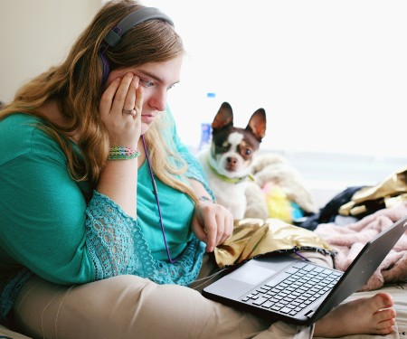 Woman wearing a teal-colored shirt, sitting on her bed wearing headphones looking at her laptop with her tiny dog at her side