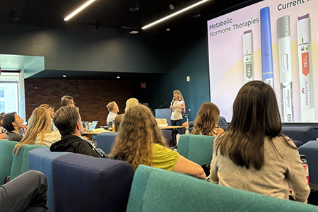 Dr. Kim Fisher is holding a microphone and presenting in front of a mixed audience. A slide behind her with a picture of metabolic hormone therapies.
