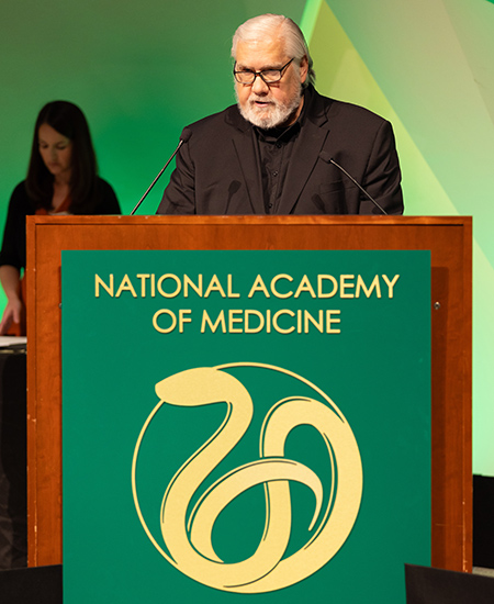 Kenneth W. Kizer is standing behind a podium with a gold print of National Academy of Medicine on green background.