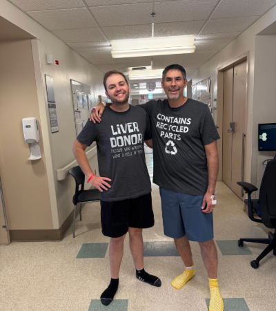 Two people in a hospital hallway wearing matching gray shirts, one labeled “Liver Donor” and the other “Contains Recycled Parts.”