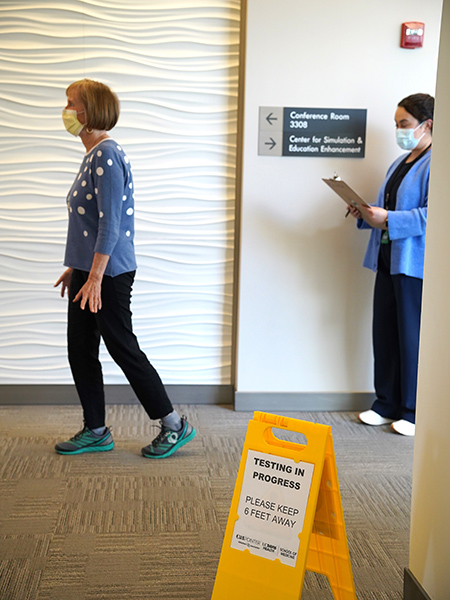 : A woman with a clipboard takes notes as an older woman practices walking in a research area designated by a yellow sign.