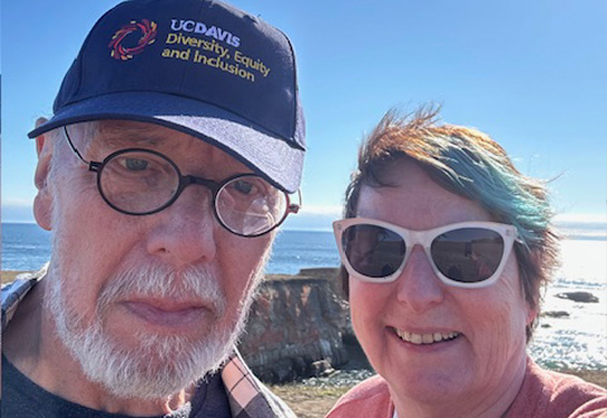 Two people smiling by the ocean, one wearing a UC Davis hat and the other white sunglasses.