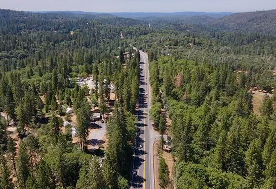 Aerial view of a rural Northern California highway surrounded by dense pine forest and scattered homes under a clear blue sky