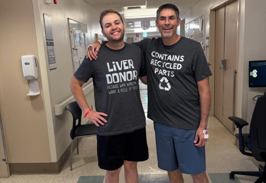 Two people in a hospital hallway wearing matching gray shirts, one labeled “Liver Donor” and the other “Contains Recycled Parts.”