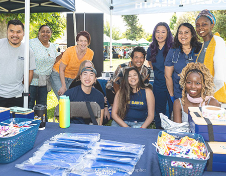 Group of people smiling at an outdoor UC Davis Health booth with tables of giveaways under a canopy at a community event