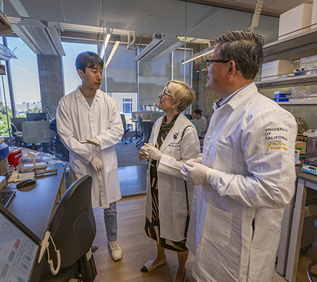 Dr. Diana Farmer standing between Dr. Aijun Wang and a researcher in Wang’s Lab. Everyone is wearing a white lab coat.
