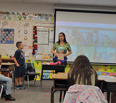 A woman stands in front of the classroom holding a doll. A boy stands to the left of her. 
