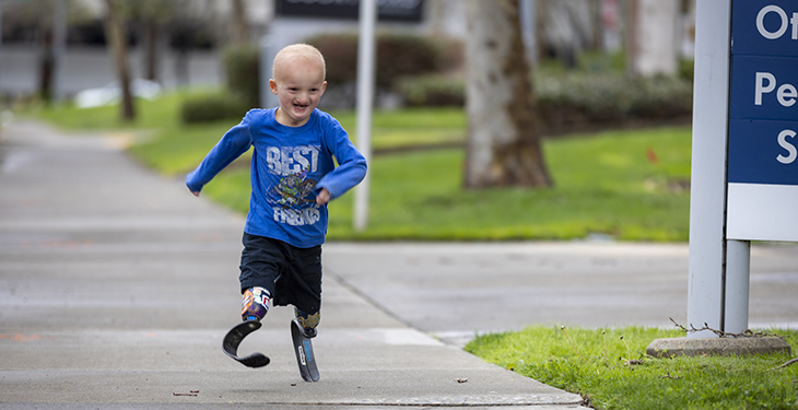 A six year old child with running blades, a blue long sleeve shirt and black shorts. He is smiling as he runs on a sidewalk.