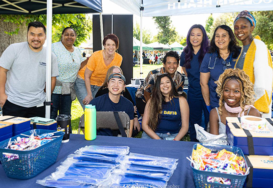 Group of people smiling at an outdoor UC Davis Health booth with tables of giveaways under a canopy at a community event