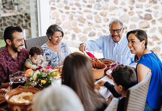 Personas sentadas a la mesa almorzando; hombre joven con bebé en brazos, tres mujeres, un niño y hombre mayor sirviendo alimento