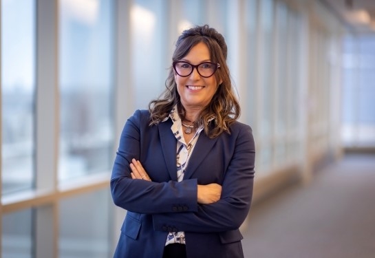 A smiling woman in glasses and a blue suit stands with arms crossed in a hallway with windows.