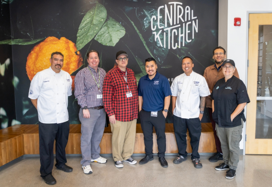 A group of seven people standing together with a “Central Kitchen” sign on the wall behind them.