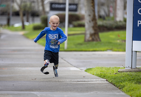: A six year old child with running blades, a blue long sleeve shirt and black shorts. He is smiling as he runs on a sidewalk.