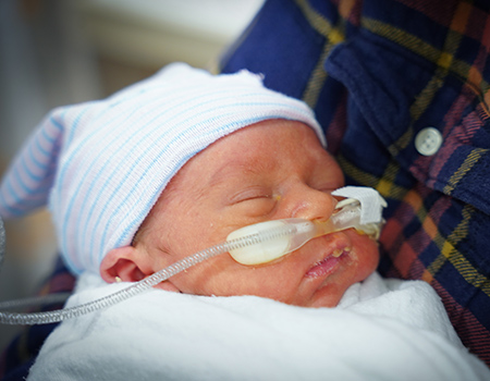 An infant with a nasal oxygen tube sleeps in a person&rsquo;s arms while wearing a white and blue stocking cap.